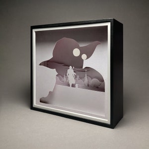 May include: A black shadow box with a white paper cutout of a character from Star Wars. The character is standing in front of a cityscape. The shadow box is on a gray background.