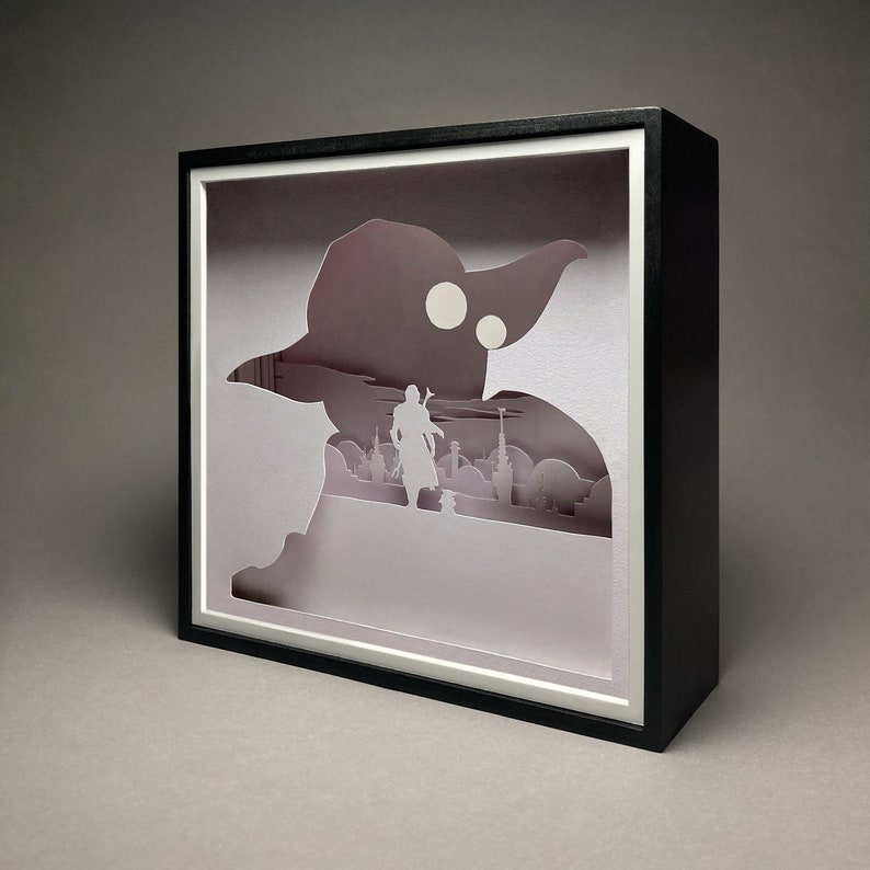 May include: A black shadow box with a white paper cutout of a character from Star Wars. The character is standing in front of a cityscape. The shadow box is on a gray background.