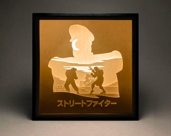 Peut inclure: Une boîte à lumière en bois noir avec une silhouette de trois personnages. Les personnages se battent devant un bâtiment. Le texte "Street Fighter" est en bas de la boîte.