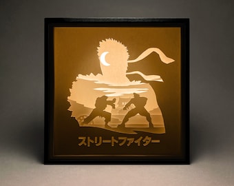 Peut inclure: Une œuvre d'art encadrée et illuminée représentant deux personnages en position de combat, avec une figure plus grande en arrière-plan. Le texte japonais "Street Fighter" est en bas. L'œuvre a un cadre noir et un intérieur chaleureux et lumineux.