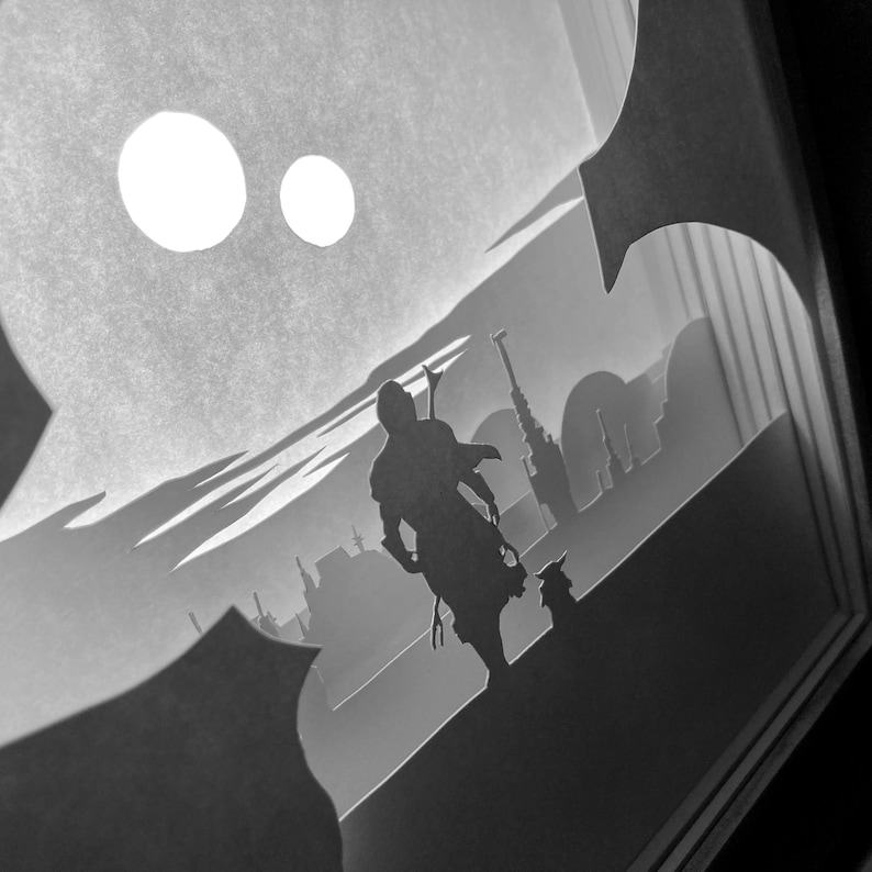 May include: A black and white papercraft scene depicting a figure with a lightsaber standing on a desert planet with two moons in the background.