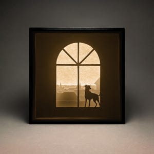 Könnte beinhalten: Ein schwarzer gerahmtes Papierkunstwerk mit einer Silhouette eines Hundes, der aus einem Fenster schaut. Das Fenster ist gewölbt und hat eine Stadtlandschaft im Hintergrund.