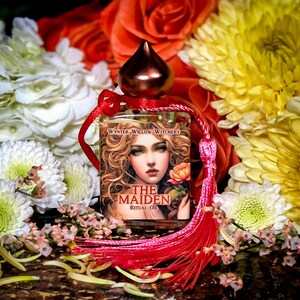 May include: A small bottle of ritual oil with a red tassel. The bottle is labeled "The Maiden Ritual Oil" and "Wynter Willow Witchery". The bottle is surrounded by white and yellow flowers.