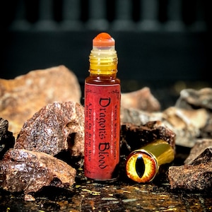 May include: A red glass bottle with a gold cap and a red label that says "Dragon's Blood" and "Winter Willow Witchery". The bottle is sitting on a brown surface with some red rocks.