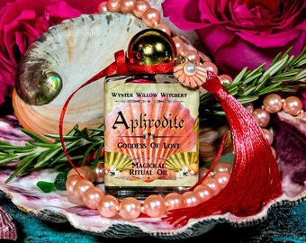 May include: A glass bottle of Aphrodite Goddess of Love Magical Ritual Oil with a red tassel and a pearl-like charm. The bottle is sitting on a seashell with pink pearls and a sprig of rosemary. The background is a dark blue surface.