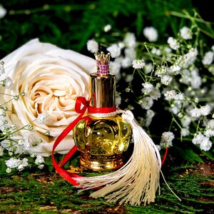 May include: A gold perfume bottle with a crown-shaped stopper and a white tassel. The bottle is surrounded by white flowers and a white rose.
