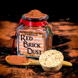 May include: A glass jar filled with red brick dust, labeled "Wynter Willow Witchery Red Brick Dust". The jar has a red ribbon tied around it and a cork lid.