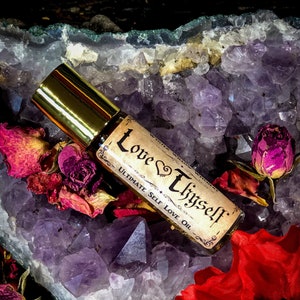 May include: A glass roller bottle with a gold cap and a label that reads "Love Yourself Ultimate Self Love Oil". The bottle is resting on a bed of purple amethyst crystals and dried rose petals.