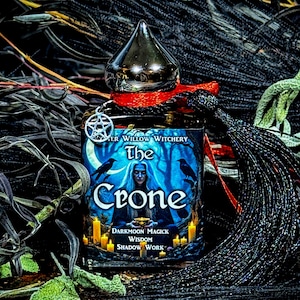 May include: A dark blue bottle with a silver top and a black tassel. The bottle has a label that says "The Crone" and "Darkmoon Magick Wisdom Shadow Work".