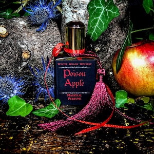 May include: A small bottle of perfume with a red tassel and a black label that reads "Poison Apple Magickal Perfume" by Wynter Willow Witchery. The bottle is sitting on a wooden surface with a red apple and green leaves.