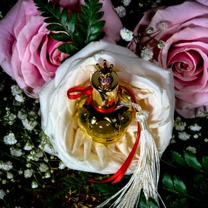 May include: A gold perfume bottle with a crown-shaped stopper and a red ribbon sits on a white rose. The bottle has a white tassel.