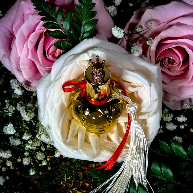 May include: A gold perfume bottle with a crown-shaped stopper and a red ribbon sits on a white rose. The bottle has a white tassel.