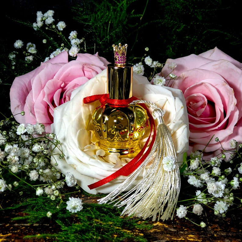 May include: A gold perfume bottle with a crown-shaped stopper and a pink gem. The bottle is surrounded by white and pink roses and small white flowers.