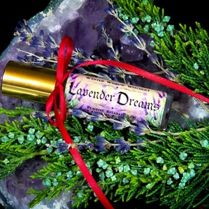 Puede incluir: Una peque&ntilde;a botella de vidrio con una tapa dorada y una cinta roja atada alrededor. La botella est&aacute; etiquetada con "Lavender Dreams" y "Winter Willow Witchery". La botella est&aacute; sentada sobre un cristal de amatista p&uacute;rpura y rodeada de follaje verde.