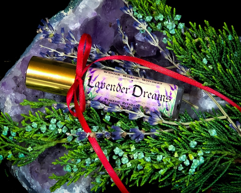 Puede incluir: Una peque&ntilde;a botella de vidrio con una tapa dorada y una cinta roja atada alrededor. La botella est&aacute; etiquetada con "Lavender Dreams" y "Winter Willow Witchery". La botella est&aacute; sentada sobre un cristal de amatista p&uacute;rpura y rodeada de follaje verde.