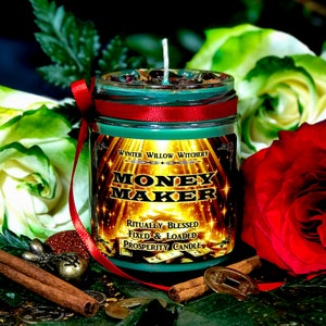 Puede incluir: Vela de vidrio verde con una cinta roja y una etiqueta dorada que dice "Money Maker" con el texto "Ritualmente Bendecido Fijo & Cargado Vela de Prosperidad" debajo. La vela est&aacute; rodeada de palitos de canela, monedas y flores.