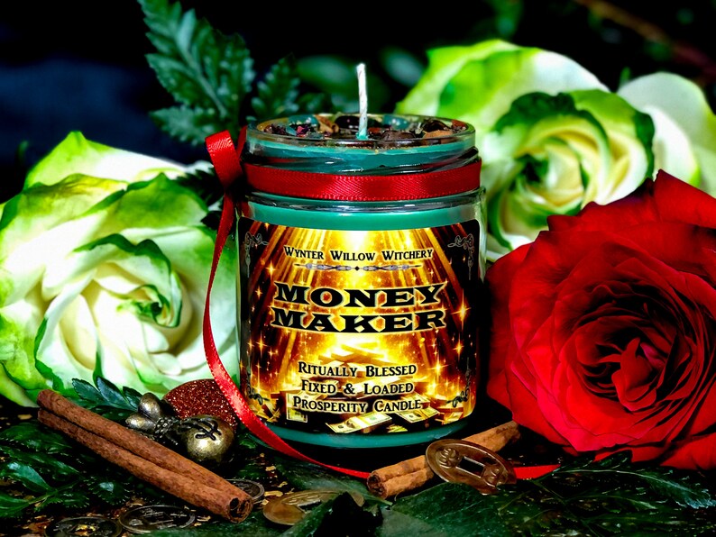 Puede incluir: Vela de vidrio verde con una cinta roja y una etiqueta dorada que dice "Money Maker" con el texto "Ritualmente Bendecido Fijo & Cargado Vela de Prosperidad" debajo. La vela est&aacute; rodeada de palitos de canela, monedas y flores.