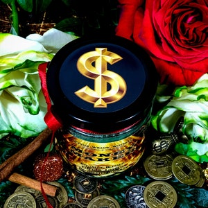 Puede incluir: Un tarro de vela negro con un signo de d&oacute;lar dorado en la tapa. El tarro est&aacute; rodeado de cinta roja, palitos de canela y monedas de oro. La vela est&aacute; etiquetada como "Money Make" y "White Willow Witchcraft".