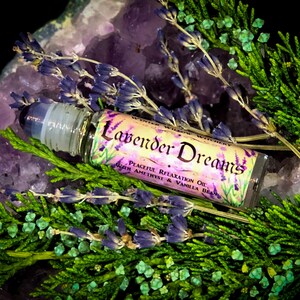 Puede incluir: Una peque&ntilde;a botella de vidrio con un aplicador de rodillo. La botella est&aacute; etiquetada "Lavender Dreams Peaceful Relaxation Oil with Amethyst & Vanilla Bean". La botella est&aacute; rodeada de cristales de amatista p&uacute;rpura y flores de lavanda.
