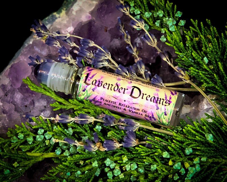 Puede incluir: Una peque&ntilde;a botella de vidrio con un aplicador de rodillo. La botella est&aacute; etiquetada "Lavender Dreams Peaceful Relaxation Oil with Amethyst & Vanilla Bean". La botella est&aacute; rodeada de cristales de amatista p&uacute;rpura y flores de lavanda.