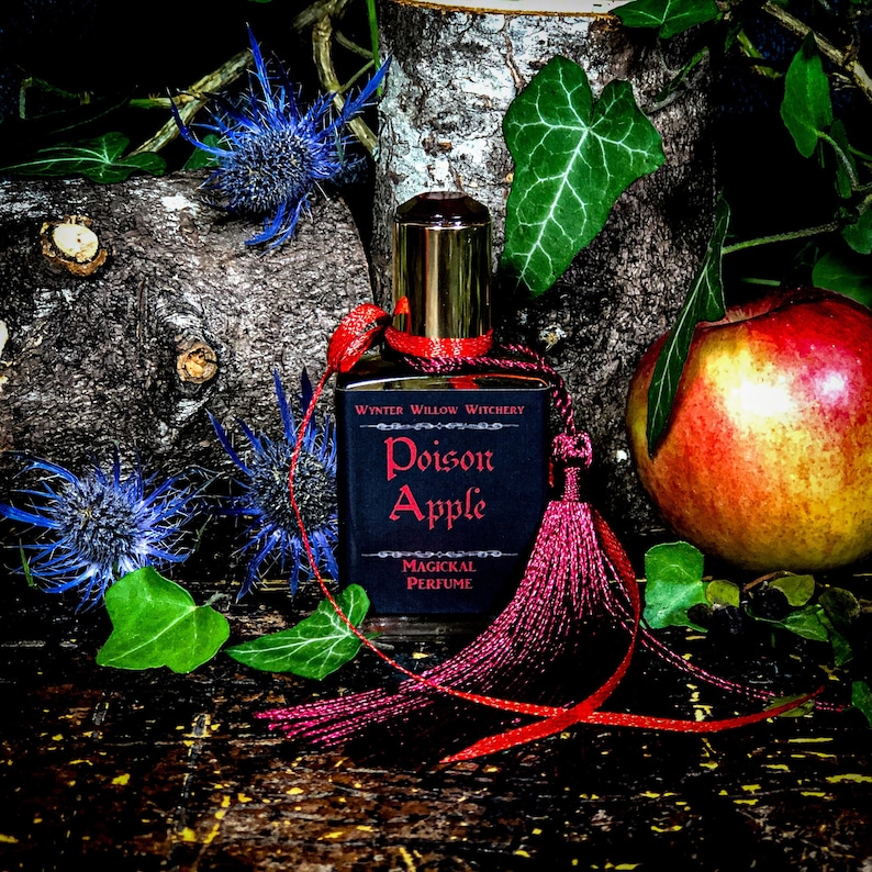 May include: A small bottle of perfume with a red tassel and a black label that reads "Poison Apple Magickal Perfume" by Wynter Willow Witchery. The bottle is sitting on a wooden surface with a red apple and green leaves.