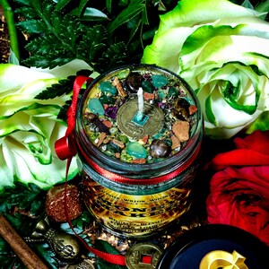 Puede incluir: Una vela en un tarro de vidrio verde con tapa roja y una etiqueta dorada que dice "Money Maker" con una cinta roja alrededor del tarro. La vela est&aacute; rodeada de hojas verdes, rosas blancas y rojas, palitos de canela y monedas de oro con caracteres chinos.