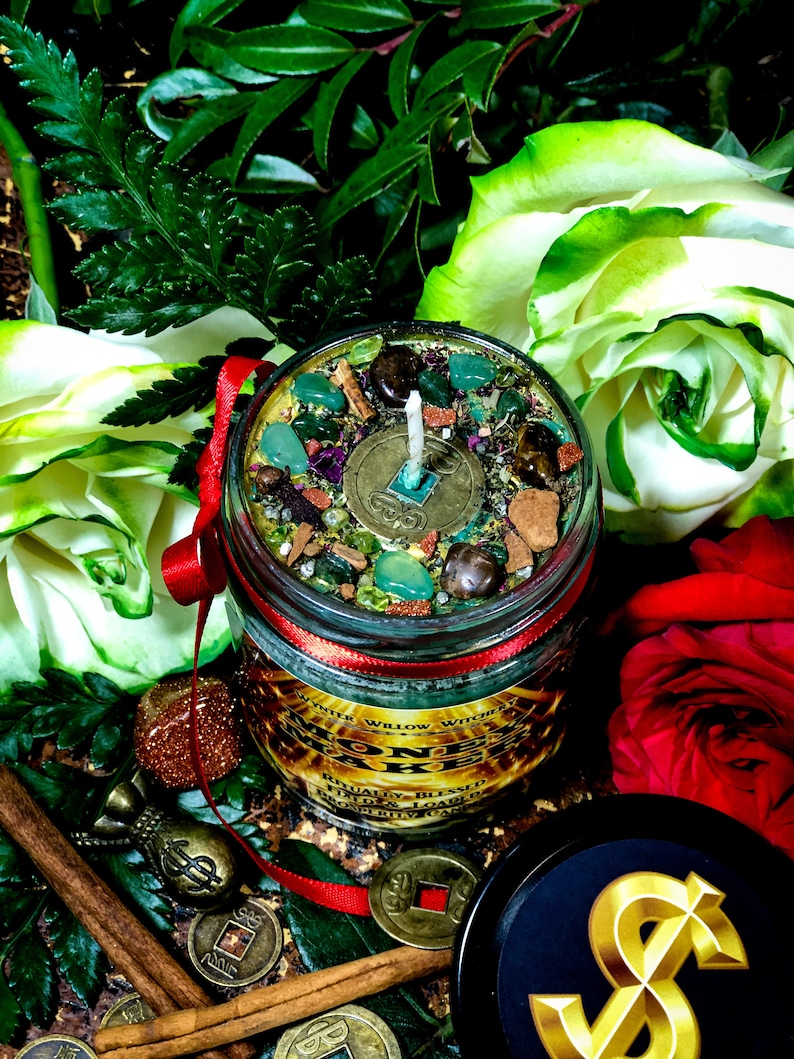 Puede incluir: Una vela en un tarro de vidrio verde con tapa roja y una etiqueta dorada que dice "Money Maker" con una cinta roja alrededor del tarro. La vela est&aacute; rodeada de hojas verdes, rosas blancas y rojas, palitos de canela y monedas de oro con caracteres chinos.