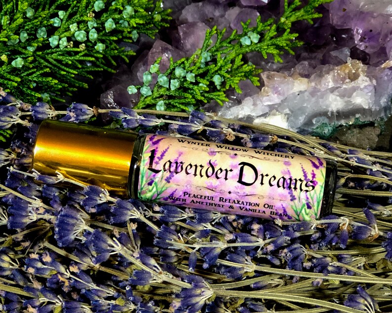 Puede incluir: Una peque&ntilde;a botella de vidrio con una tapa dorada y una etiqueta que dice "Lavender Dreams Peaceful Relaxation Oil with Amethyst & Vanilla Bean". La botella est&aacute; rodeada de flores de lavanda y cristales de amatista.