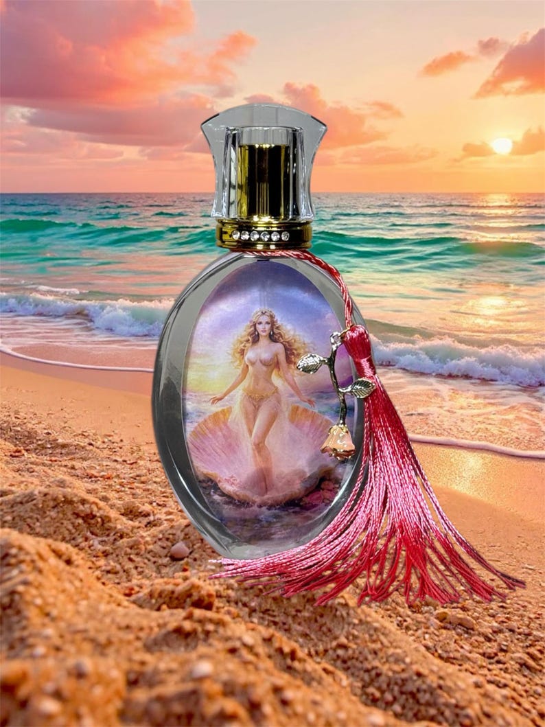 Puede incluir: Frasco de perfume ovalado con cuerpo de vidrio transparente y tap&oacute;n dorado. El frasco presenta una imagen de una mujer en una concha. Una borla rosa y una rosa decorativa acent&uacute;an el frasco. El fondo muestra una escena de playa con una puesta de sol.