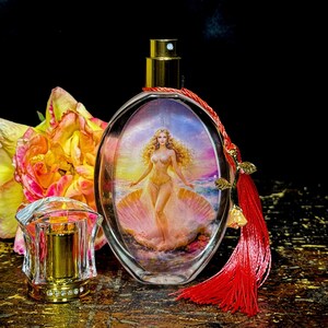 Puede incluir: Frasco de perfume ovalado con tap&oacute;n dorado y pintura de mujer en concha. La botella tiene borla roja y est&aacute; junto a tap&oacute;n decorativo transparente y flor amarilla y naranja.