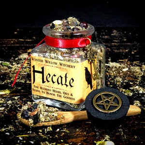 May include: A glass jar with a red ribbon and a label that reads "WYNTER WILLOW WITCHERY Hecate FINE BLENDED HERBS, OILS & RESINS TO HONOR THE GODDESS". The jar is filled with dried herbs and other ingredients. A wooden spoon with some of the ingredients on it is next to the jar. A black charcoal disc with a gold pentagram on top is also in the image.