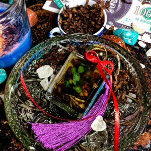 May include: A clear glass bottle with a red ribbon and a purple tassel. The bottle is filled with a green liquid and contains small green and brown pieces. The bottle is sitting on a glass plate with a decorative pattern. There are also some small white crystals and brown tobacco leaves on the plate.