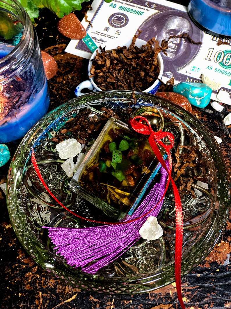 May include: A clear glass bottle with a red ribbon and a purple tassel. The bottle is filled with a green liquid and contains small green and brown pieces. The bottle is sitting on a glass plate with a decorative pattern. There are also some small white crystals and brown tobacco leaves on the plate.
