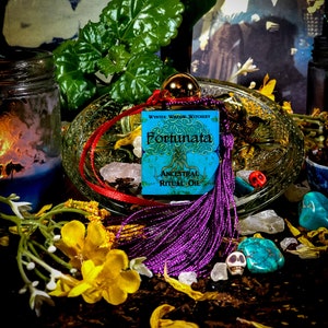 May include: A small blue bottle with a purple tassel and a label that reads "Wynter Willow Witchery Fortunata Ancestral Ritual Oil". The bottle is sitting on a glass dish with crystals and flowers.