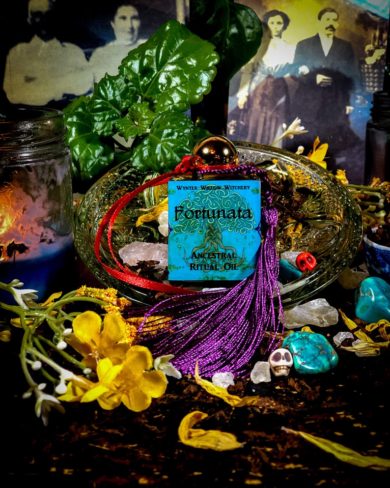 May include: A small blue bottle with a purple tassel and a label that reads "Wynter Willow Witchery Fortunata Ancestral Ritual Oil". The bottle is sitting on a glass dish with crystals and flowers.