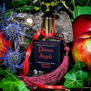 May include: A glass bottle of Poison Apple magickal perfume with a red tassel and a black label. The bottle is surrounded by green leaves, red apples, and blue thistle flowers.