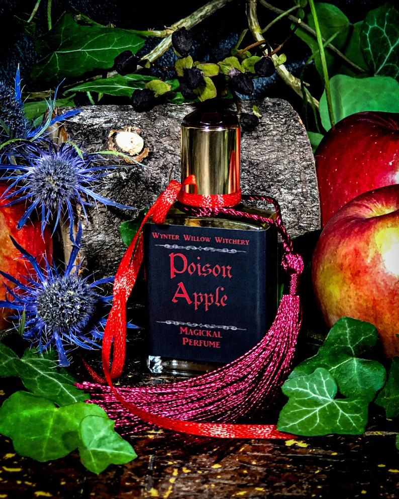 May include: A glass bottle of Poison Apple magickal perfume with a red tassel and a black label. The bottle is surrounded by green leaves, red apples, and blue thistle flowers.