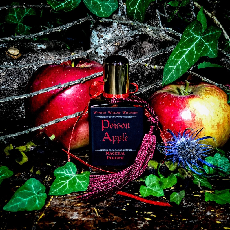 May include: A dark glass bottle with a gold cap and a red tassel. The bottle is labeled "Poison Apple" and "Magickal Perfume". The bottle is surrounded by red apples and green leaves.