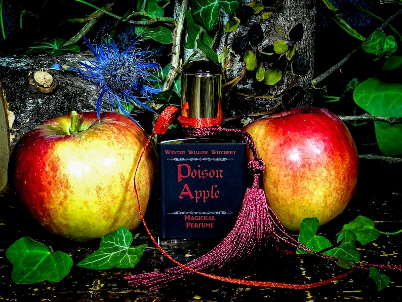 May include: A dark glass bottle with a gold top and a red tassel. The bottle is labeled "Winter Willow Witchery Poison Apple Magickal Perfume". Two red apples are in the background.