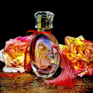 Puede incluir: Un frasco de perfume ovalado con un tap&oacute;n transparente y facetado. El frasco presenta una imagen pintada de una mujer y est&aacute; adornado con una cinta y una borla rojas. El fondo incluye rosas coloridas.