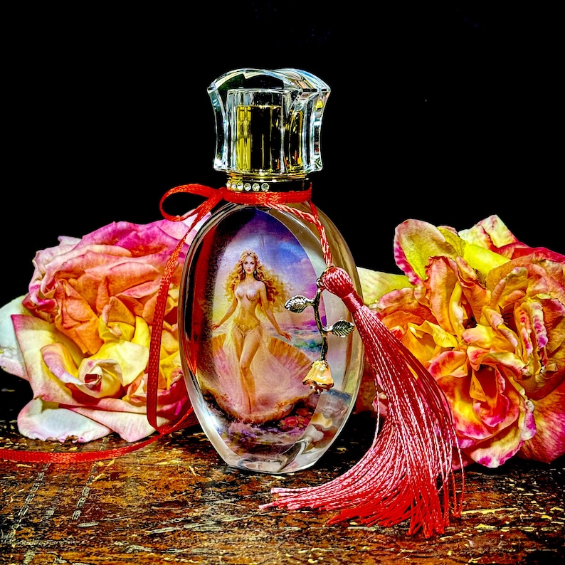 Puede incluir: Un frasco de perfume ovalado con un tap&oacute;n transparente y facetado. El frasco presenta una imagen pintada de una mujer y est&aacute; adornado con una cinta y una borla rojas. El fondo incluye rosas coloridas.