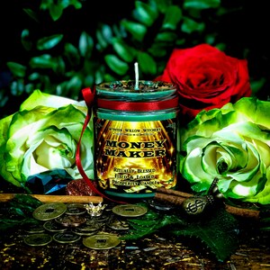 Puede incluir: Una vela verde y dorada con el texto "Money Maker" y "Ritually Blessed Fixed & Loaded Prosperity Candle" en la etiqueta. La vela est&aacute; rodeada de hojas verdes, monedas y una rama de canela.