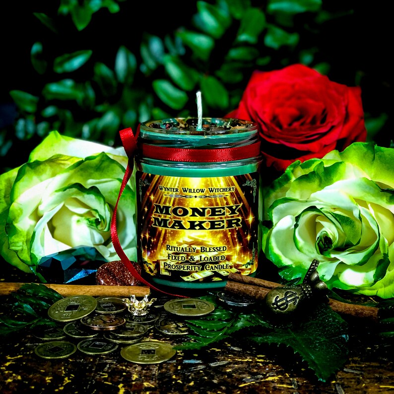 Puede incluir: Una vela verde y dorada con el texto "Money Maker" y "Ritually Blessed Fixed & Loaded Prosperity Candle" en la etiqueta. La vela est&aacute; rodeada de hojas verdes, monedas y una rama de canela.