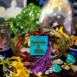 May include: A small blue bottle with a purple tassel and a label that reads "Wynter Willow Witchery Fortunata Ancestral Ritual Oil". The bottle is surrounded by various crystals, flowers, and other witchy items.