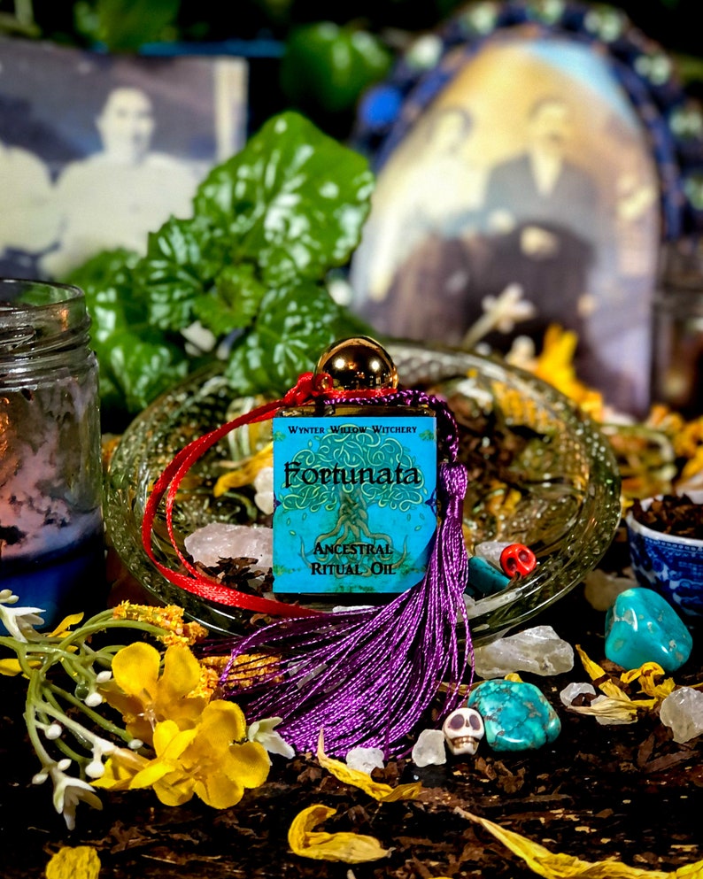 May include: A small blue bottle with a purple tassel and a label that reads "Wynter Willow Witchery Fortunata Ancestral Ritual Oil". The bottle is surrounded by various crystals, flowers, and other witchy items.