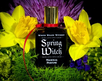 May include: A black bottle of perfume with a gold cap and a red ribbon. The bottle is labeled "Spring Witch Magickal Perfume" and is surrounded by yellow daffodils and purple flowers.