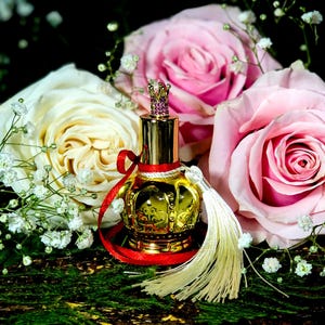 May include: A gold perfume bottle with a crown-shaped stopper and a red ribbon. The bottle is surrounded by white and pink roses and baby's breath.