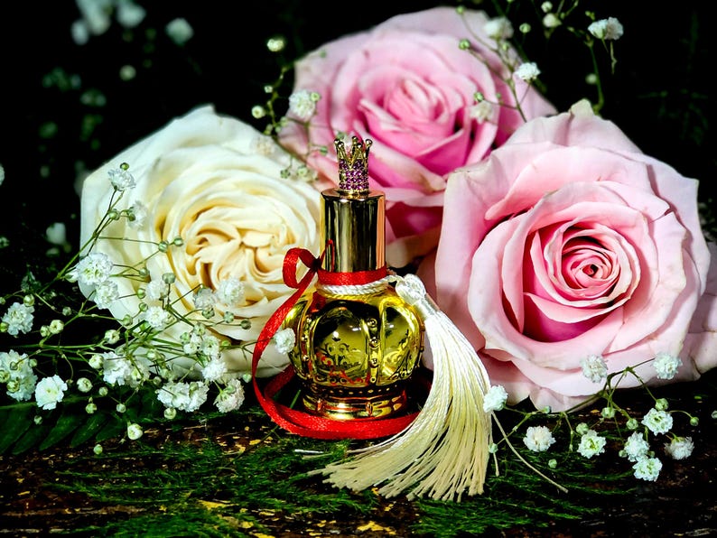 May include: A gold perfume bottle with a crown-shaped stopper and a red ribbon. The bottle is surrounded by white and pink roses and baby's breath.