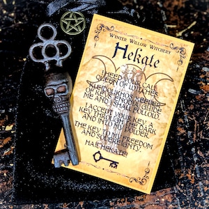 May include: A black velvet bag with a key pendant, a key-shaped charm, and a gold-colored pentagram charm. A card with the text "Hekate" and a poem is also included. The key pendant is shaped like a skull.