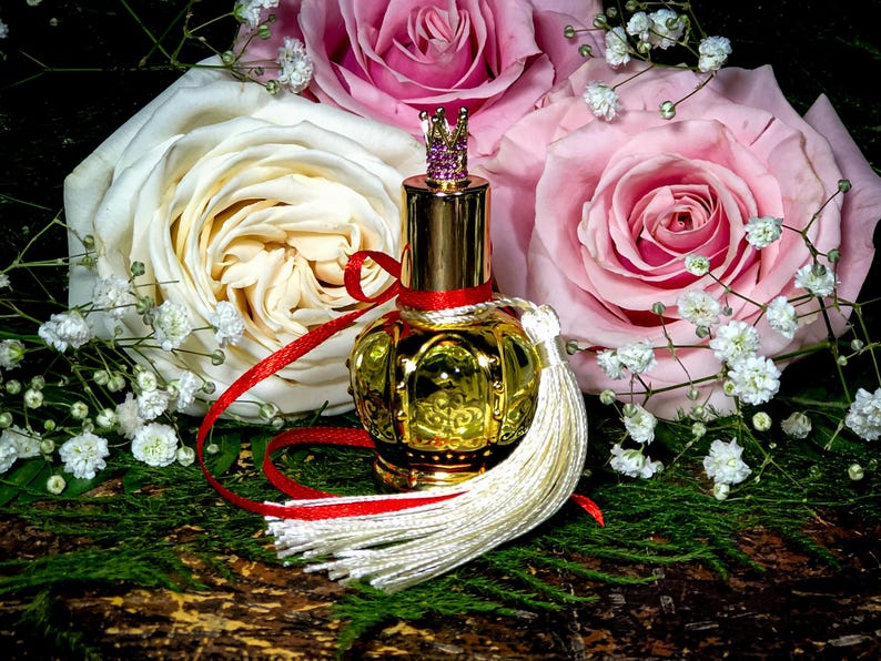 May include: A gold perfume bottle with a crown-shaped stopper and a white tassel, surrounded by pink and white roses and baby's breath.